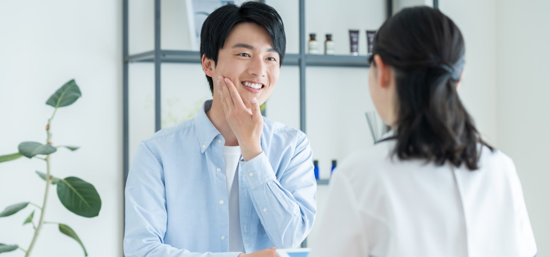 頬に手を当てて笑顔を見せる男性と、後ろで見守る女性。棚にはスキンケア製品が並ぶ清潔な空間